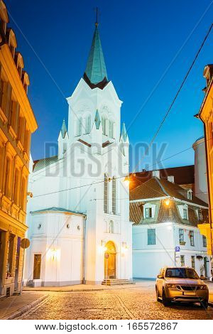 Riga Latvia. The White Tower With Pyramidal Spire Of Our Lady Of Sorrows Or Virgin Of Anguish Church, Ancient Catholic Church On Pils Street In Evening Illumination Under Summer Blue Sky