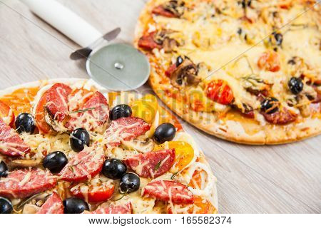 Pizza cutter together with two large pizzas with cheese olives and salami mushrooms and tomatoes. On a wooden stand.