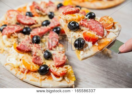 Cook takes a shovel one piece of pizza with cheese olives and salami mushrooms and tomatoes. On a wooden stand.