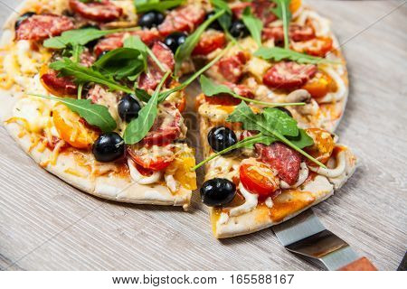 Cook takes a shovel one piece of pizza with cheese olives and salami mushrooms and tomatoes arugula. On a wooden stand.