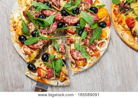Cook takes a shovel one piece of pizza with cheese olives and salami mushrooms and tomatoes arugula. On a wooden stand.