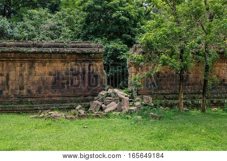 Stone wall of an ancient ruin with a big gap from a break through. Loose stone blocks from the breach can be seen on the ground. The wall is part of the ancient ruins of Angkor.