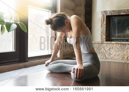 Young attractive woman practicing yoga, sitting in Uddiyana Bandha exercise, Upward Abdominal Lock pose, working out, wearing sportswear, grey pants, bra, indoor full length, home interior background