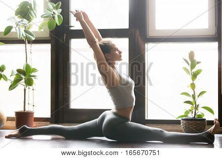 Young attractive woman practicing yoga, sitting in Monkey God, Splits exercise, Hanumanasana pose, working out, wearing sportswear, grey pants, bra, indoor full length, home interior background