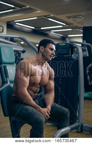 Young bodybuilder looking at the muscles after a workout. Handsome guy sitting on the simulator. Bodybuilder tired after a workout. The man sitting in the gym. A guy with a naked torso. Sexy guy.