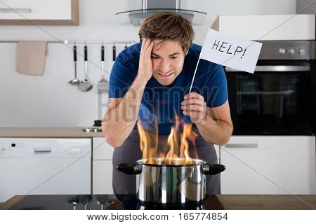 Worried Young Man Holding Help Flag With Utensil On Fire On Electric Stove At Home