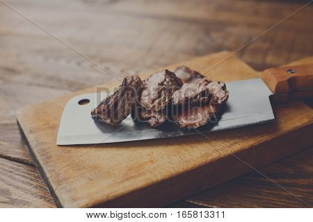Cleaver butcher's knife slicing grilled steak on meat cutting board on wooden background. Fresh juicy roasted meat cuts. Chef cooking