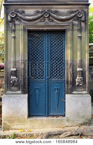 Blue steel door with rectangular frame as entrance to a crypt / tomb. It is ornate with a cross pattern, floral decorations and big vases. The doorframe contains chiseled reliefs of torches carved into the stone.