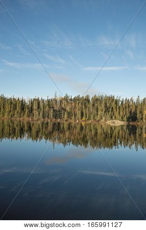 Calm lake in early morning with reflections of conifers and wispy cirrus clouds