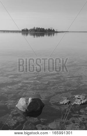 Dead calm afternoon image of shallow and clear lake huron water and limestone rocks along theshoreline. A small tree-covered island is on the horizon. Overall feeling is a calm peaceful tranquil serene and wild background in this black and white capture.