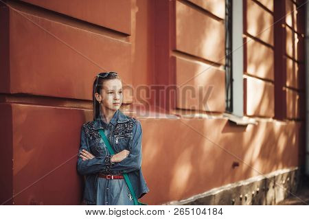 Serious Little Girl In Jeans Clothes Stands Near An Old Restored Beautiful Building.