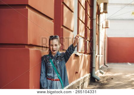 Cheerful Little Girl In Jeans Clothes Stands Near An Old Restored Beautiful Building.