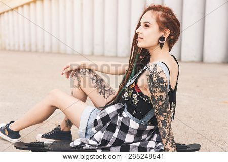 Young Girl With Tattoo And Dreadlocks Sitting On A Longboard On The Background Of Concrete Structure