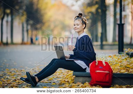 Pretty Hipster Teen Girl Sitting On A Sidewalk On Autumn City Street And Working Laptop Computer. Sc
