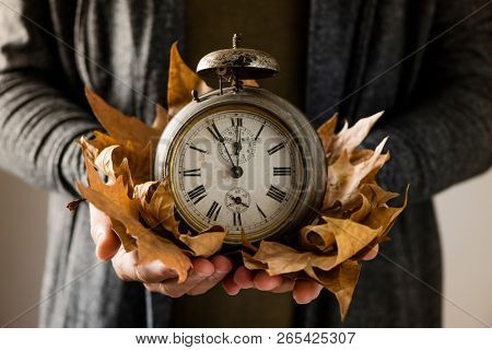 closeup of a young man with an old and rusty alarm clock surrounded by dry leaves in his hands, depicting the end of the summer time and the beginning of autumn