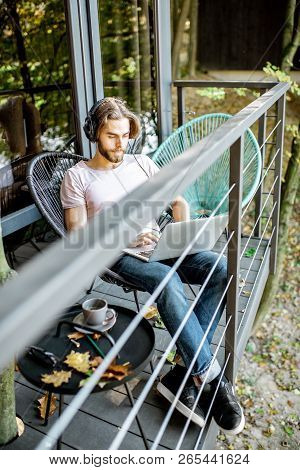 Poortrait Of A Handsome Man Working With Laptop And Headphones On The Balcony Of The House In The Be