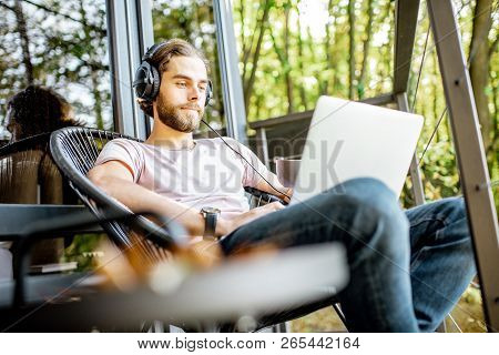 Poortrait Of A Handsome Man Working With Laptop And Headphones On The Balcony Of The House In The Be