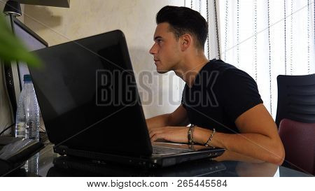 Attractive Young Man With Serious Expression, With Laptop And Desktop Comptuers, Very Busy Working O