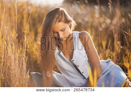 Beautiful Young Woman In Jean Sundress Sitting Relxed In A Field Of Tall Grass On Sunset And Reading
