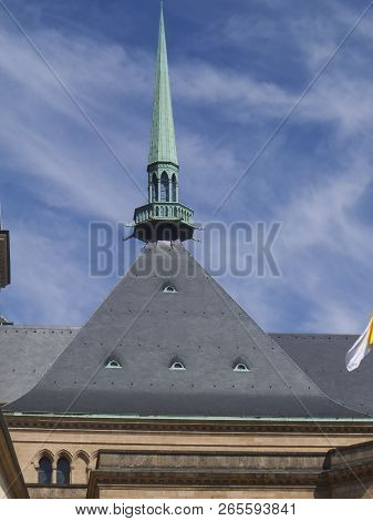 A Roof Top In Luxembourg. A Nice Grey Roof With A Spike On Top In Green And The Blue Sky In The Back