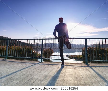 Runner Doing Stretching Exercise On Bay Bridge. An Active Wiry Man Standing On One Legs And Stretchi