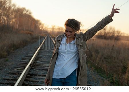 Young Attractive Lady In White Shirt And Beige Raincoat Walking Along The Railroad Tracks Around Blu