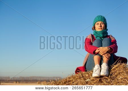 Young Attractive Lady In Blue Knitting Hat And Pink Coat Sitting On Haystack. Smiling Beautiful Hips
