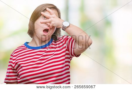 Young adult woman with down syndrome over isolated background peeking in shock covering face and eyes with hand, looking through fingers with embarrassed expression.