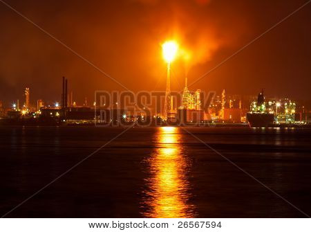 Oil refinery at night creating a huge smoke cloud with reflections on the nearby ocean