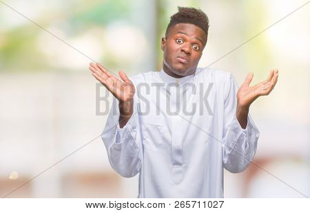 Young arabic african man wearing traditional djellaba over isolated background clueless and confused expression with arms and hands raised. Doubt concept.