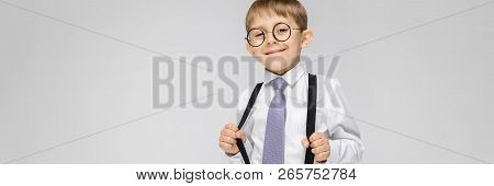 A Charming Boy In A White Shirt, Suspenders, A Tie And Light Jeans Stands On A Gray Background. The 
