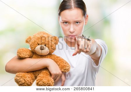 Young caucasian woman holding teddy bear over isolated background pointing with finger to the camera and to you, hand sign, positive and confident gesture from the front
