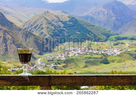 A Glass Of Georgian Red Wine Stands On A Wooden Railing On The Background Of The Georgian Village An