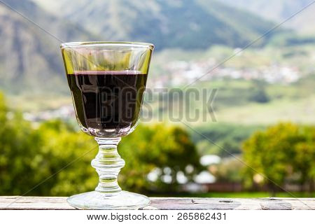 A Glass Of Georgian Red Wine Stands On A Wooden Railing On The Background Of The Georgian Village An