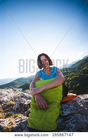 A Woman Is Sitting In A Sleeping Bag At Sunset. Camping On Top Of A Mountain. A Smiling Girl Rests A