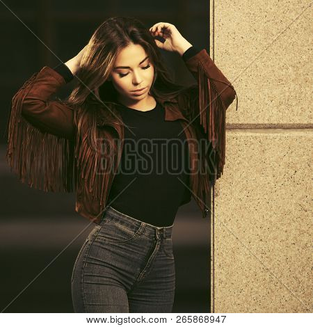 Young woman with long straight hairs walking in city street  Stylish fashion model wearing brown leather fringe suede jacket and dark blue jeans