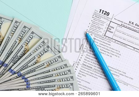 1120 Tax Form Lies Near Hundred Dollar Bills And Blue Pen On A Light Blue Background. Us Corporation