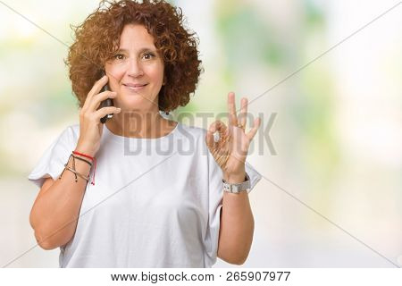Middle ager senior woman talking on smartphone over isolated background doing ok sign with fingers, excellent symbol
