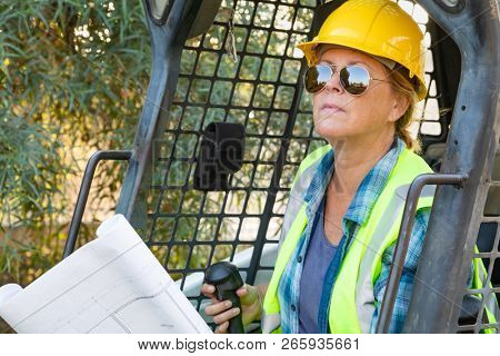 Smiling Female Worker Holding Technical Blueprints Using Small Bulldozer At Constrcution Site