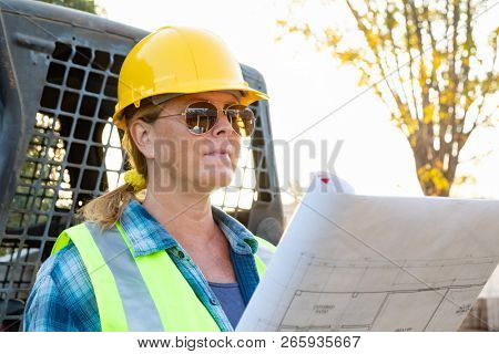 Female Worker Holding Technical Blueprints Near Small Bulldozer At Constrcution Site