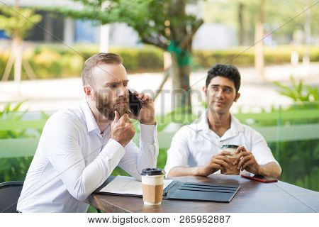 Thoughtful Businessman Rubbing Beard While Talking On Phone In Outdoor Cafe. Frowning Male Manager T