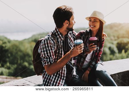 Happy Couple Sitting On Ledge And Drinking Coffee. Young Man And Woman With Backpacks Sitting Togeth