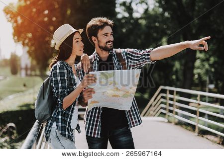 Young Smiling Couple Looking At Map Outdoors. Casualy Dressed Happy Travelers With Backpacks Plannin