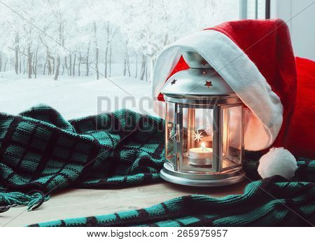 Festive tranquil Christmas and New Year still life -lantern with candle and Christmas Santa hat on the windowsill and winter park outdoor. Concept of Christmas and New Year.Christmas and New year still life. Christmas composition