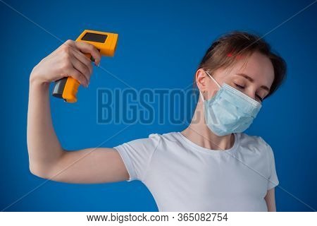 Woman In Medical Face Mask Holding Yellow Pyrometer And Showing Suicide Gesture Like Pointing Gun To