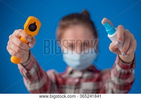 Woman Holding Yellow Pyrometer And Spraying Antiseptic Toward Camera Against Blue Wall - Close Up, S