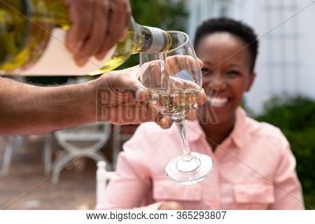 Senior African American woman and her family spending time together in the garden on a sunny day, a man is pouring wine into a glass in the foreground.