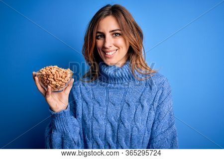 Young beautiful brunette woman holding bowl with peanuts over blue background with a happy face standing and smiling with a confident smile showing teeth