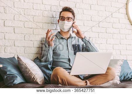 Happy Hipster Man Freelancer In Medical Face Mask Talking Mobile Phone, Sitting On Couch With Laptop