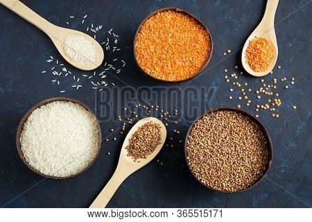 Three Full Bowls Of Cereals Lentils, Rice And Buckwheat On A Dark Blue Background. The Wooden Spoons